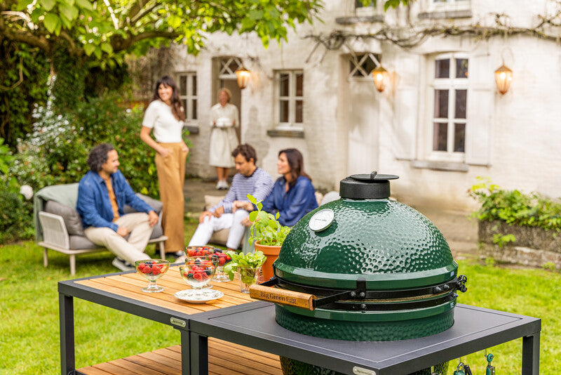 Ein Big Green Egg im Garten, daneben Schälchen mit Beeren und Basilikum in Töpfen, dahinter eine Familie auf der Lounge