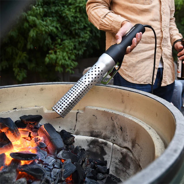 Person using a handheld electric fire starter over a fire pit with glowing coals.