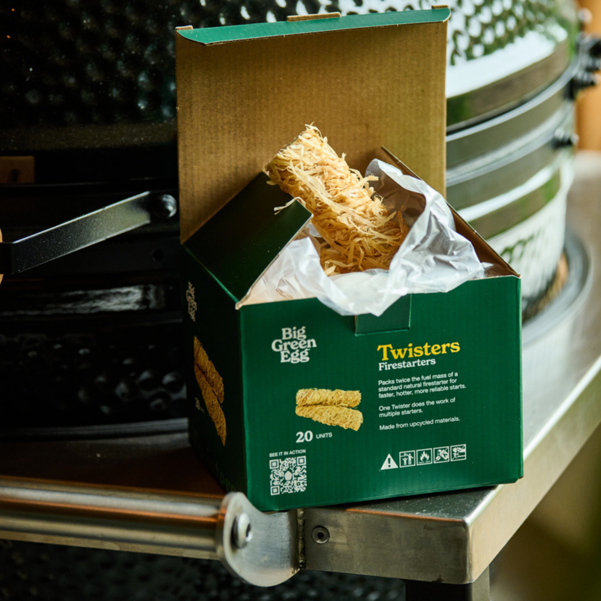 A green box of Big Green Egg Twisters Firestarters containing a bundle of natural fiber firestarter rods, shown open on a metal surface in front of a grill.