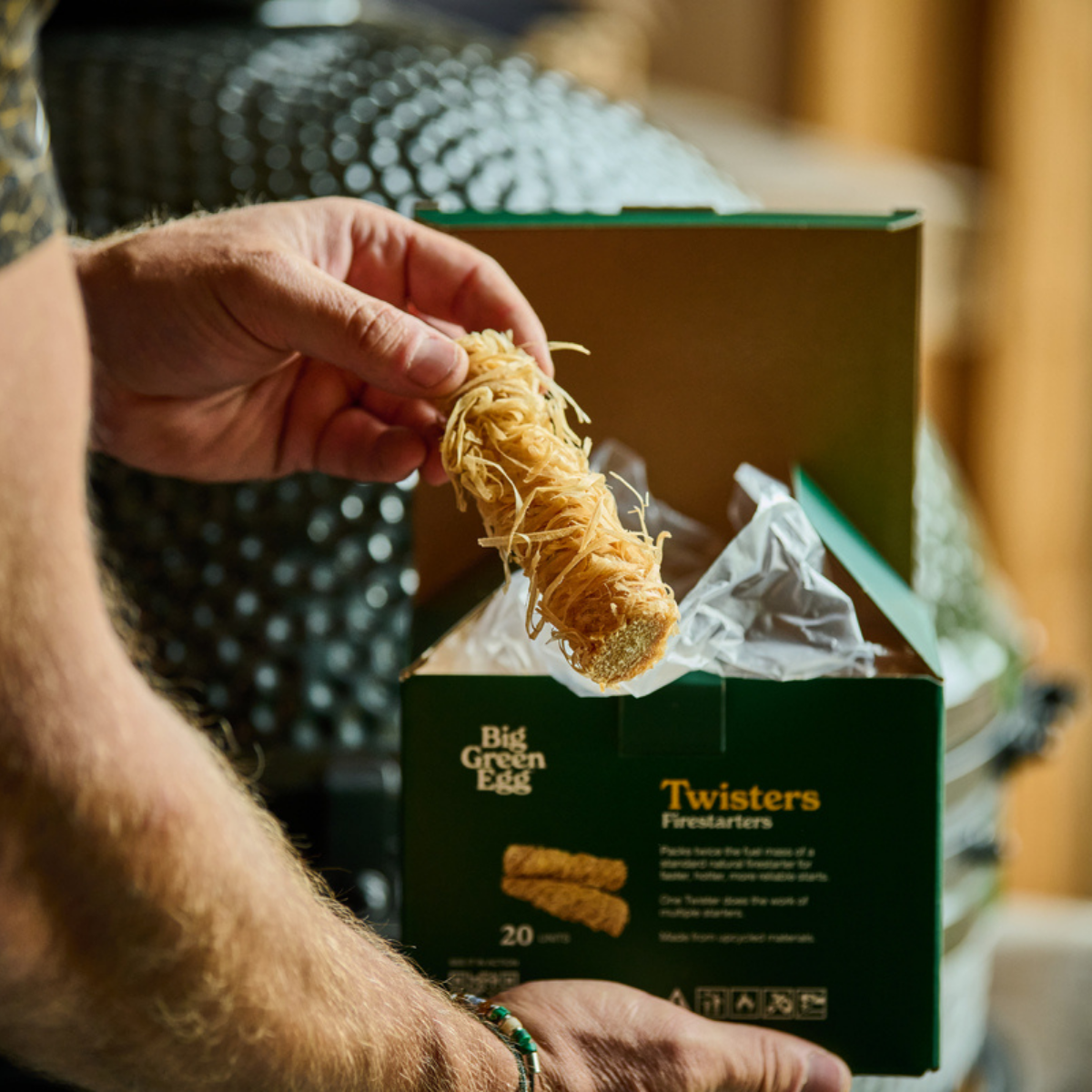A person's hands hold a Twister firestarter stick above an open green box labeled Big Green Egg Twisters Firestarters, with shredded paper filler inside the box.