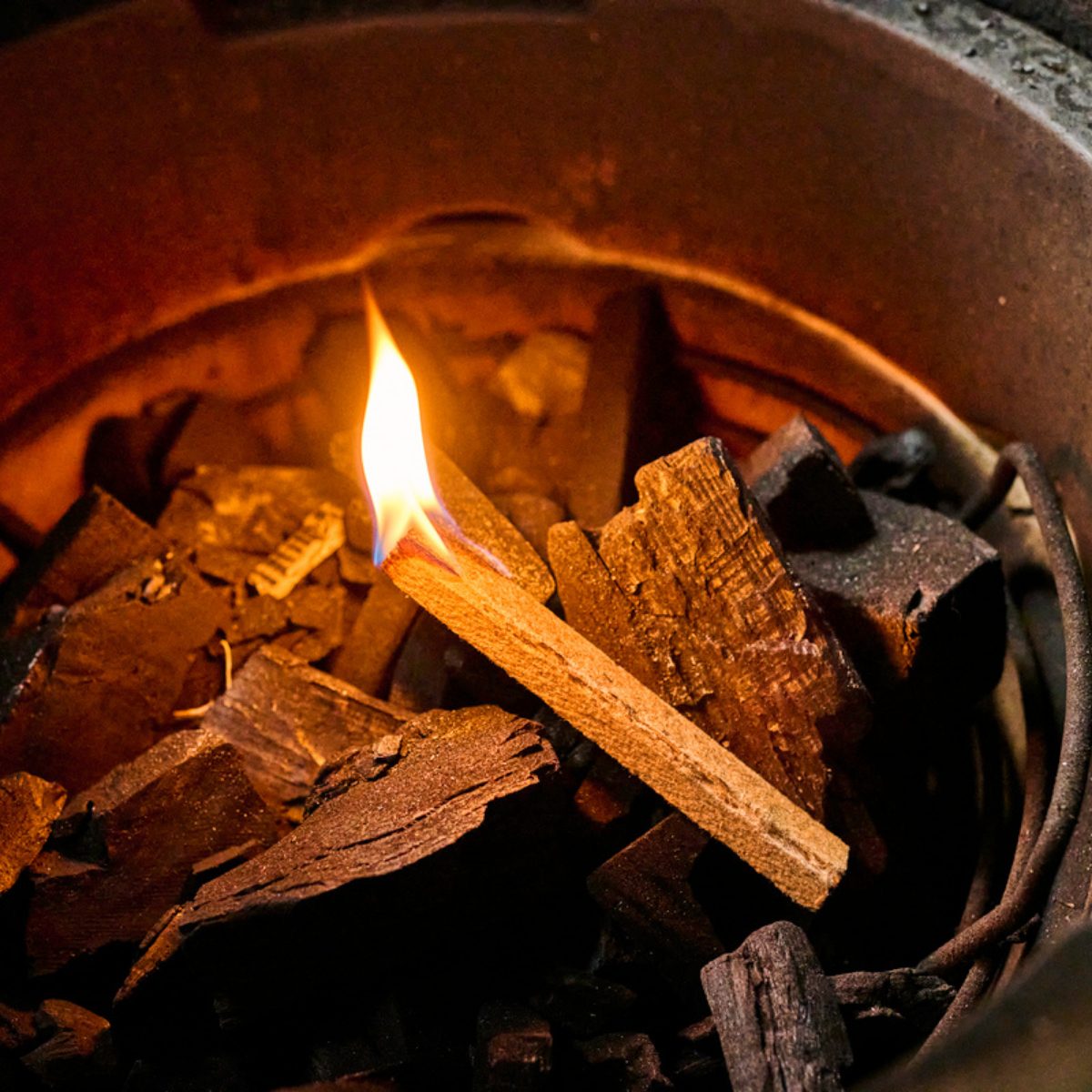 A close-up of a SpeediLight charcoal starter lighting dark charcoal coals inside a Big Green Egg, with a small flame lit and warm orange light reflecting off the wood textures.