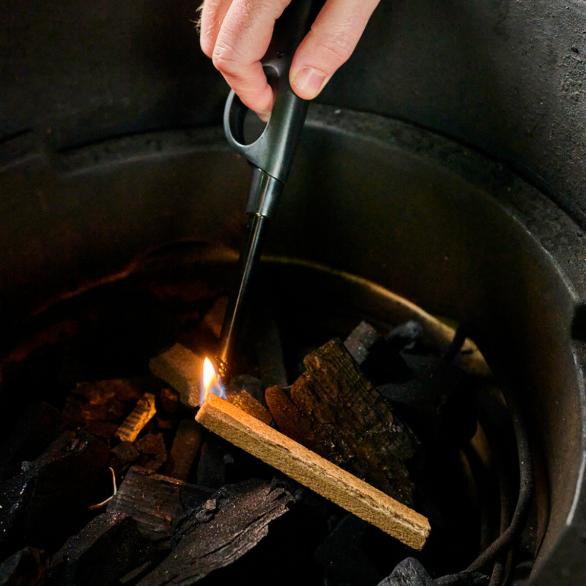 A close-up of a hand holding a black long-reach lighter and lighting a stick of Speedilight charcoal starter inside of a Big Green Egg.