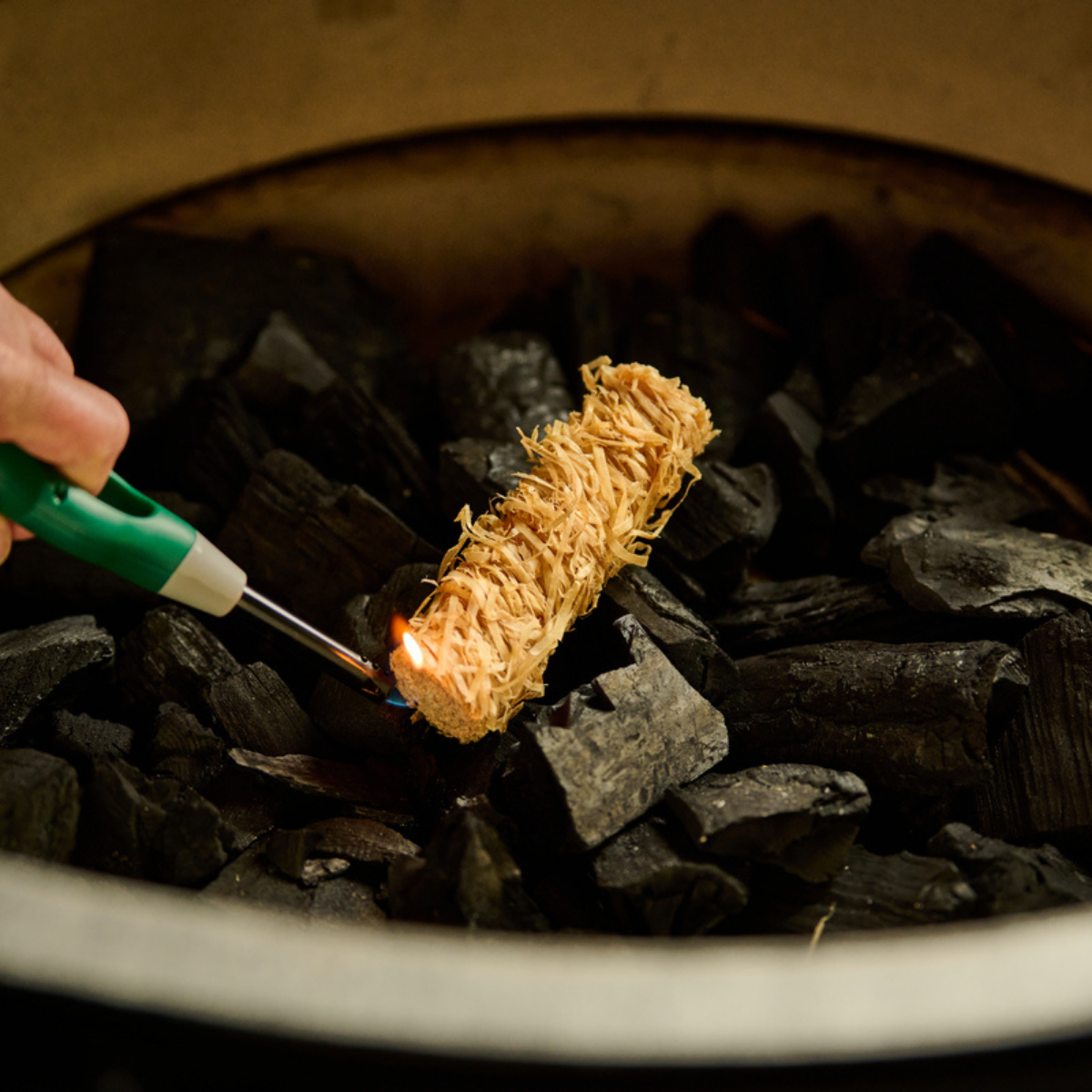 A hand holds a green and white lighter to ignite a bundle of natural fiber starter wood over black charcoal in a fire pit.
