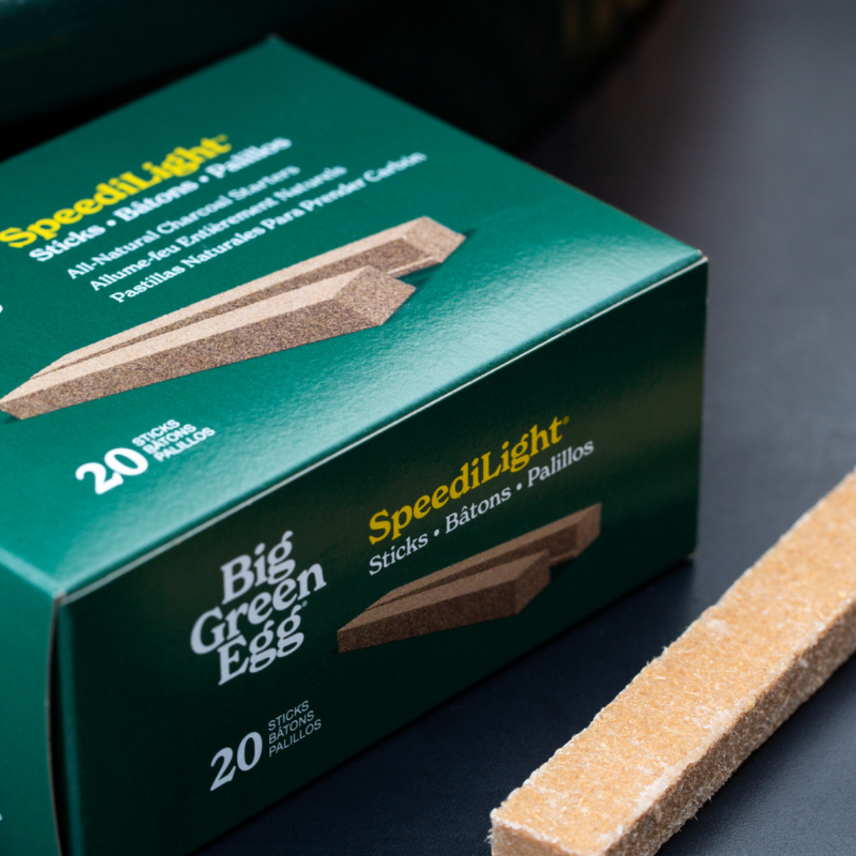 A close-up of a green SpeediLight Big Green Egg charcoal starter stick packaging with a single natural wood stick placed diagonally in the foreground on a dark surface.